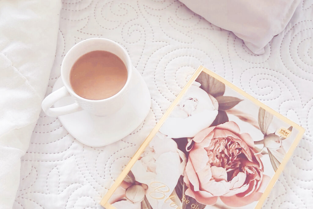 Fragment łóżka. Biała narzuta. Na niej leży notes z okładką w kwiaty.  Obok stoi kubek z kawą
