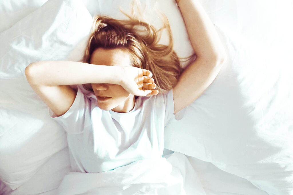 Kobieta. Leży w łóżku. Jedną ręką zakrywa oczy. Druga ręka uniesiona jest nad głową.