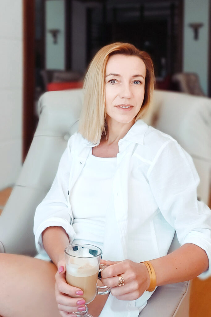 kobieta. Blond włos. Siedzi na beżowym fotelu. W ręku trzyma szklankę z kawą.