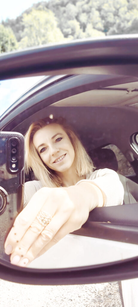 Zdjęcie odbicia w lusterku bocznym samochodu. Kobieta trzyma smartfon, którym robi zdjęcie. 