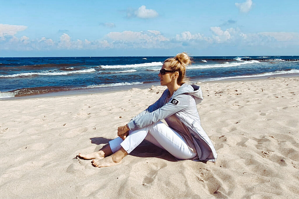 Kobieta siedzi na plaży. Ubrana w białe spodnie, szarą kurtkę. Włosy spięte w kok. Patrzy w dal, w stronę morza.